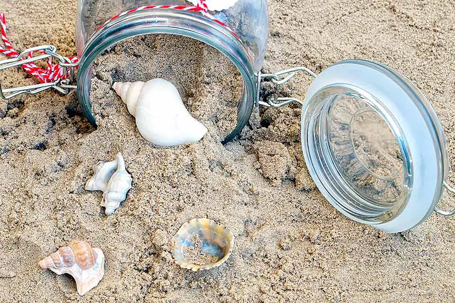 Jar laying in sand, half filled with sand and assorted sea shells