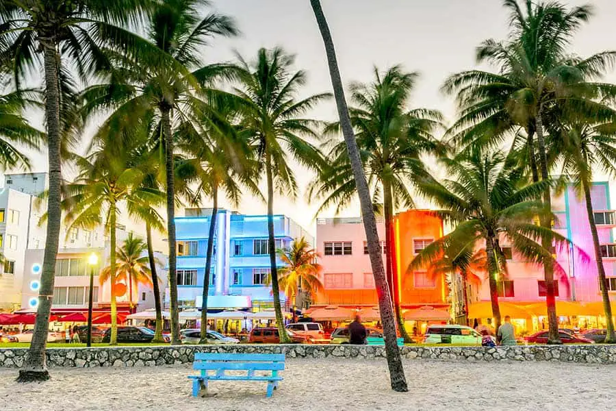 Palm trees, blue bench on sandy beach and colorful buildings in the background