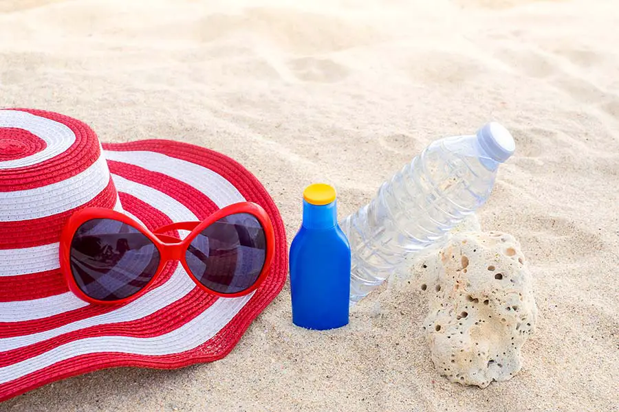 Red and white sunhat, sunglasses, sunblock and water laying on beach sand